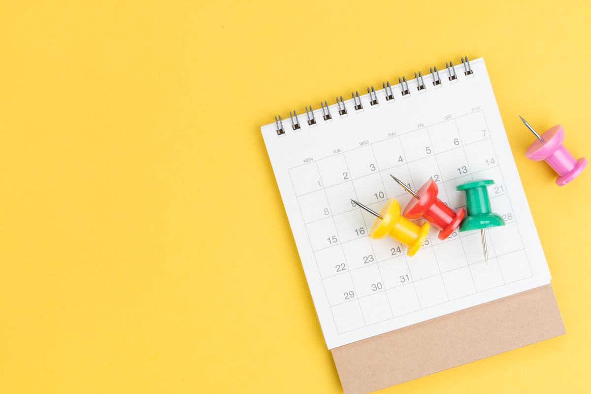 Flat lay or top view of colorful oversized thumbtack or pushpin on white calendar on yellow background, appointment, reminder, schedule and date organizer concept